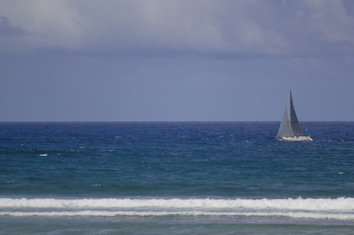 A sailboat is moving across a vast expanse of deep blue ocean under a cloudy sky. The water is slightly choppy, creating small waves near the shoreline, and the horizon is visible in the distance.