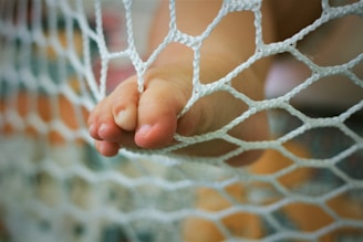 A small foot with toes pressed against a white netting material. The focus is on the toes and net, with a blurred background that includes soft colors.