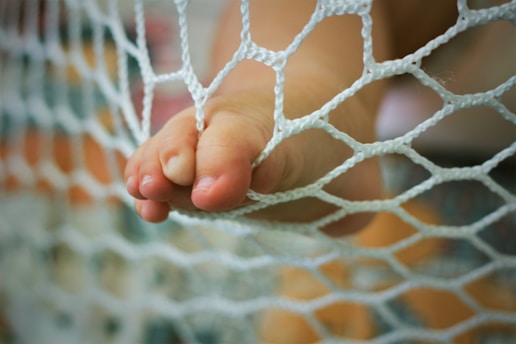 A small foot with toes pressed against a white netting material. The focus is on the toes and net, with a blurred background that includes soft colors.