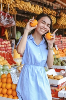 A woman in a blue dress is smiling and holding an orange in each hand. She is standing in front of a colorful fruit stand filled with bananas, oranges, and other fruits.