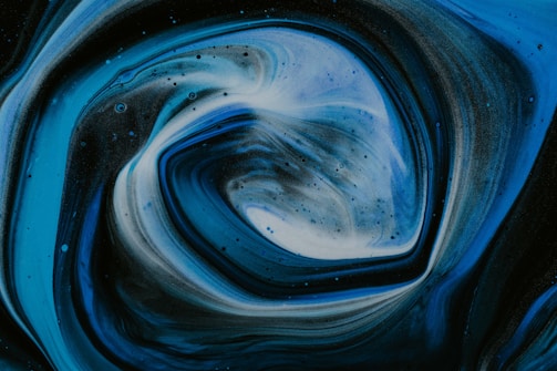 Swirling patterns create a mesmerizing vortex effect with gradients of blue, black, and white. The texture resembles a fluid mixture, capturing a sense of motion and depth.