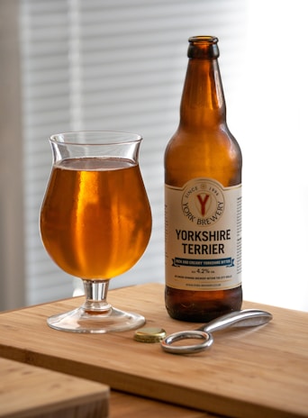 A glass of amber-colored beer is placed next to a bottle labeled 'Yorkshire Terrier' from York Brewery. Both are on a wooden surface, accompanied by a bottle opener and a bottle cap, with a softly lit background featuring horizontal blinds.