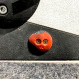 A close-up view of a rock climbing hold with two screws that resemble eyes, mounted on a textured climbing wall. The hold is orange and shaped somewhat like a rounded triangle. The wall has a combination of black and white sections.