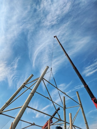 A large crane is assembling a metal structure under a clear blue sky with scattered clouds. The structure consists of long metal beams arranged at various angles, forming a geometric framework. Workers can be seen operating the crane and managing the construction process.