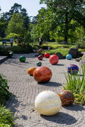 A serene garden setting with a series of colorful spherical sculptures arranged on a gravel path. The background features lush greenery and several people standing or sitting on benches, enjoying the calm atmosphere.