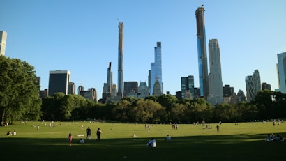 A large green park with people scattered across the grass, enjoying leisure activities. Towering skyscrapers dominate the background skyline under a clear blue sky. Trees border the open space, adding a natural element to the urban scene.