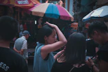 A group of people are gathered in a vibrant marketplace, with colorful umbrellas overhead. The main focus is on a woman adjusting her hair, while another person beside her is looking at a phone. Shop stalls display various items in the background, creating a busy atmosphere.