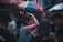A group of people are gathered in a vibrant marketplace, with colorful umbrellas overhead. The main focus is on a woman adjusting her hair, while another person beside her is looking at a phone. Shop stalls display various items in the background, creating a busy atmosphere.