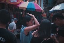A group of people are gathered in a vibrant marketplace, with colorful umbrellas overhead. The main focus is on a woman adjusting her hair, while another person beside her is looking at a phone. Shop stalls display various items in the background, creating a busy atmosphere.