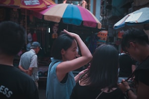 A group of people are gathered in a vibrant marketplace, with colorful umbrellas overhead. The main focus is on a woman adjusting her hair, while another person beside her is looking at a phone. Shop stalls display various items in the background, creating a busy atmosphere.