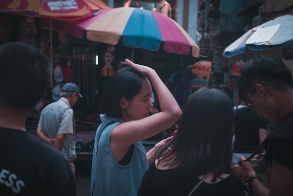 A group of people are gathered in a vibrant marketplace, with colorful umbrellas overhead. The main focus is on a woman adjusting her hair, while another person beside her is looking at a phone. Shop stalls display various items in the background, creating a busy atmosphere.