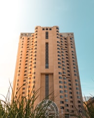 A tall beige building with a rectangular structure, featuring numerous windows and a large vertical column of glass windows in the center. In the foreground, there are green plants partially covering the lower part of the building. The InterContinental logo is visible near the top left side of the structure. The sky in the background is clear and blue with sunlight illuminating the scene.