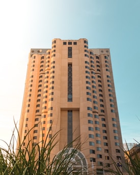 A tall beige building with a rectangular structure, featuring numerous windows and a large vertical column of glass windows in the center. In the foreground, there are green plants partially covering the lower part of the building. The InterContinental logo is visible near the top left side of the structure. The sky in the background is clear and blue with sunlight illuminating the scene.