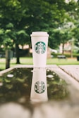 A Starbucks coffee cup is placed on a wooden surface outdoors, with a reflection visible in a puddle of water. The background is filled with lush green trees, providing a natural and calming atmosphere.