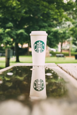 A Starbucks coffee cup is placed on a wooden surface outdoors, with a reflection visible in a puddle of water. The background is filled with lush green trees, providing a natural and calming atmosphere.