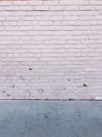 A textured brick wall painted white with visible cracks and some peeling areas. The ground is concrete with visible cracks and stains.