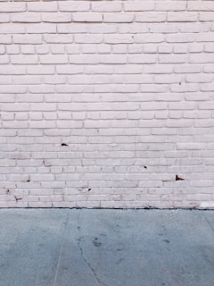 A textured brick wall painted white with visible cracks and some peeling areas. The ground is concrete with visible cracks and stains.