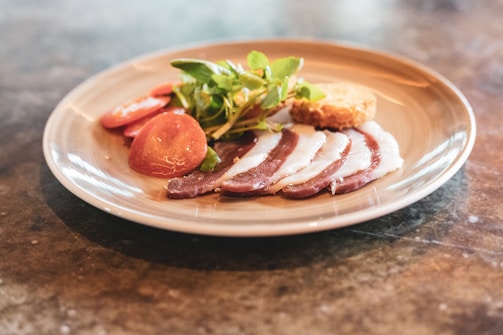 A plate with slices of cured meat, fresh green salad, slices of tomato, and a piece of crusty bread is placed on a marble countertop.