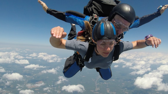 Two people are tandem skydiving with one person harnessed to a professional instructor. They are free-falling through a clear blue sky, with fluffy white clouds below them. Both individuals are wearing helmets and protective eyewear, and the instructor is holding onto the harness for stability.