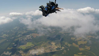 A tandem skydiving pair freefalling over a vibrant patchwork of farmland under clear blue skies.
