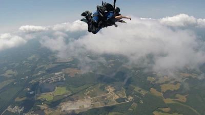 A tandem skydiving pair freefalling over a vibrant patchwork of farmland under clear blue skies.