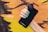 A hand with turquoise nail polish holds a black smartphone with a large camera lens attachment, against a background of a colorful, graffiti-covered brick wall.