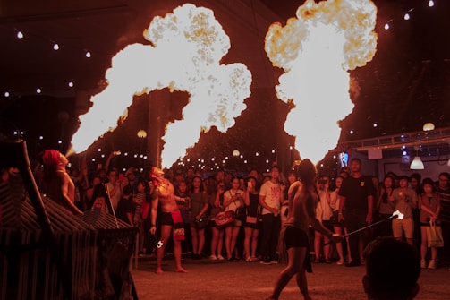 A crowd gathers to watch fire performers in an evening setting. The performers, scantily clad, are in mid-action, breathing large flames into the air. String lights hang above, adding to the festive atmosphere.