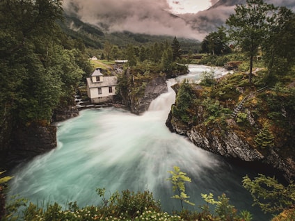A house perched on the edge of a rocky riverbank surrounded by lush green trees. The river flows rapidly, creating a dynamic and serene natural scene. The background features misty mountains partially obscured by clouds.