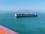 A large cargo ship carrying numerous colorful shipping containers is sailing on a calm greenish-blue sea. In the background, a mountainous landscape is visible under a clear blue sky. The image is partially framed by an orange-colored foreground, possibly a railing.