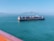 A large cargo ship carrying numerous colorful shipping containers is sailing on a calm greenish-blue sea. In the background, a mountainous landscape is visible under a clear blue sky. The image is partially framed by an orange-colored foreground, possibly a railing.
