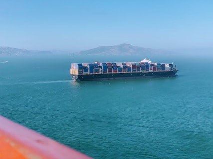 A large cargo ship carrying numerous colorful shipping containers is sailing on a calm greenish-blue sea. In the background, a mountainous landscape is visible under a clear blue sky. The image is partially framed by an orange-colored foreground, possibly a railing.
