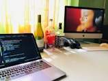 On a desk, there is an open laptop displaying code on its screen. Next to the laptop, several bottles, including wine and whiskey, are lined up. An iMac with a blurred image as wallpaper, a keyboard, mouse, and a game controller are also present. Sunlight streams through a window in the background, creating a warm ambiance.