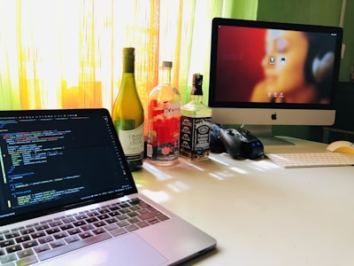 On a desk, there is an open laptop displaying code on its screen. Next to the laptop, several bottles, including wine and whiskey, are lined up. An iMac with a blurred image as wallpaper, a keyboard, mouse, and a game controller are also present. Sunlight streams through a window in the background, creating a warm ambiance.