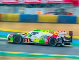 A racing car with vibrant graffiti-style art on its body zooms along a race track. The background includes colorful barriers and blurred elements, indicating motion and speed.
