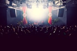 A crowded nightclub scene featuring a DJ booth illuminated by bright lights. The crowd is silhouetted against the stage, where DJs are performing. Colorful banners hang from the ceiling, and there are large speakers visible on both sides. Some palm plants add a tropical atmosphere to the setting.