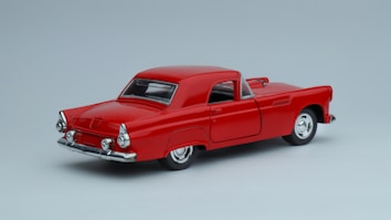 A classic red vintage car model is positioned against a plain light gray background. The car features a sleek, shiny surface with chrome accents on the bumpers and hubcaps. Its design highlights round tail lights and elegant curves typical of mid-20th-century automobiles.