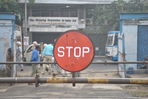 A red circular sign with the word 'STOP' in white letters is prominently displayed in the foreground, mounted on a horizontal metal bar. In the background, there are several people walking near a building with a sign that reads 'The Shipping Corporation of India Ltd.' A blue and white vehicle is parked on the right side near blue structures. The scene is set outdoors with partial views of greenery.