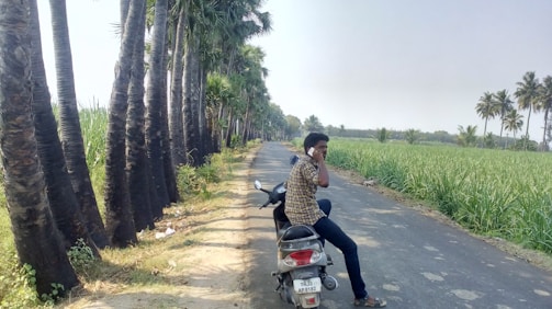 A friendly customer service agent holding a phone with Bali motorbikes in the background under tropical palm trees.