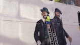 A street artist wearing a yellow safety vest is painting a large mural on a white brick wall. The mural depicts two gentlemen in vintage attire, including suits and hats. One gentleman is dressed in a green coat and bowler hat, and the other in a darker outfit with a beret. The artist stands on a ladder, working on the details of the painting.