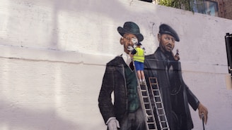 A street artist wearing a yellow safety vest is painting a large mural on a white brick wall. The mural depicts two gentlemen in vintage attire, including suits and hats. One gentleman is dressed in a green coat and bowler hat, and the other in a darker outfit with a beret. The artist stands on a ladder, working on the details of the painting.