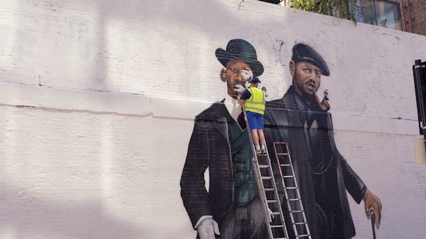 A street artist wearing a yellow safety vest is painting a large mural on a white brick wall. The mural depicts two gentlemen in vintage attire, including suits and hats. One gentleman is dressed in a green coat and bowler hat, and the other in a darker outfit with a beret. The artist stands on a ladder, working on the details of the painting.