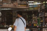 A man in a light blue shirt stands in front of a busy shop counter filled with various boxed products. The shelves are densely packed with items, mostly in rectangular packaging. The shop appears to offer a wide range of products, possibly including cigarettes, over-the-counter medicines, and other general goods.