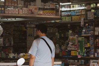 A man in a light blue shirt stands in front of a busy shop counter filled with various boxed products. The shelves are densely packed with items, mostly in rectangular packaging. The shop appears to offer a wide range of products, possibly including cigarettes, over-the-counter medicines, and other general goods.