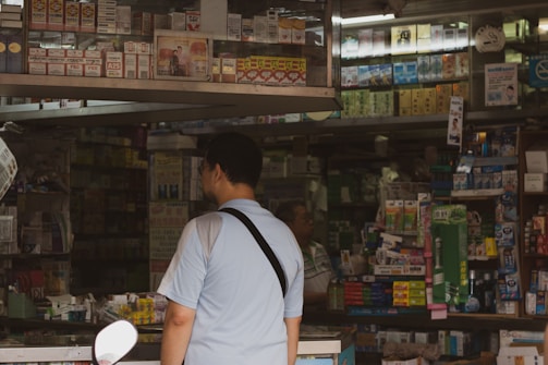A man in a light blue shirt stands in front of a busy shop counter filled with various boxed products. The shelves are densely packed with items, mostly in rectangular packaging. The shop appears to offer a wide range of products, possibly including cigarettes, over-the-counter medicines, and other general goods.