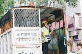 A person wearing a yellow safety helmet and reflective vest stands on the open back of a truck. The truck features metal mesh on the sides and appears to be used for transportation or delivery. Another person, partially visible, is approaching the truck from the side. The background includes trees and building signage with various writings.