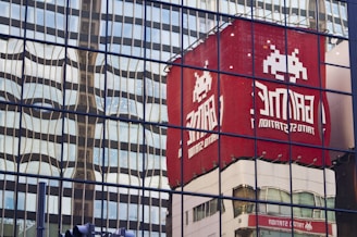 Reflections of a red building featuring a pixel art alien character and the text 'GAME' are visible on a glass facade. The mirrored surface creates a wavy distortion effect, merging the reflection with the geometric patterns of the glass panels.