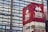 Reflections of a red building featuring a pixel art alien character and the text 'GAME' are visible on a glass facade. The mirrored surface creates a wavy distortion effect, merging the reflection with the geometric patterns of the glass panels.