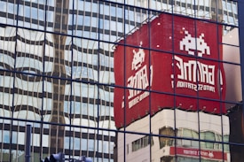 Reflections of a red building featuring a pixel art alien character and the text 'GAME' are visible on a glass facade. The mirrored surface creates a wavy distortion effect, merging the reflection with the geometric patterns of the glass panels.