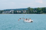 A group of people are on a small motorboat in the middle of a large body of water. One person is diving off the side of the boat into the water while others observe. In the background, there are buildings surrounded by trees along the shoreline.