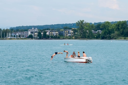 A group of people are on a small motorboat in the middle of a large body of water. One person is diving off the side of the boat into the water while others observe. In the background, there are buildings surrounded by trees along the shoreline.